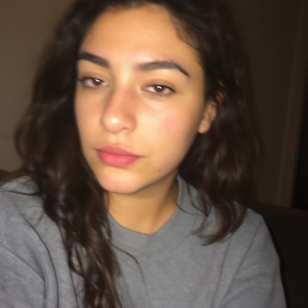 Mujer de cabello largo y oscuro aparece en una selfie algo desenfocada. Lleva sudadera gris y mira a la cámara con expresión seria y relajada.